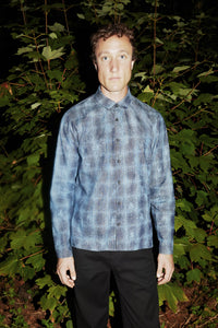 A man standing outdoors at night in front of dense green foliage, wearing a long-sleeve denim button-up shirt with a gray plaid pattern that seems pencil drawn and black pants.