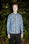 A man standing outdoors at night in front of dense green foliage, wearing a long-sleeve denim button-up shirt with a gray plaid pattern that seems pencil drawn and black pants.