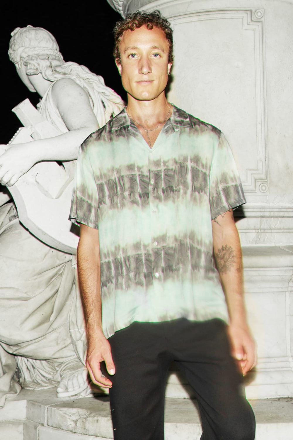 A man wearing a short-sleeve button-up shirt with a light green and gray tie-dye pattern, paired with black pants, standing in front of a white stone statue at night.