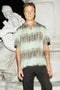 A man wearing a short-sleeve button-up shirt with a light green and gray tie-dye pattern, paired with black pants, standing in front of a white stone statue at night.
