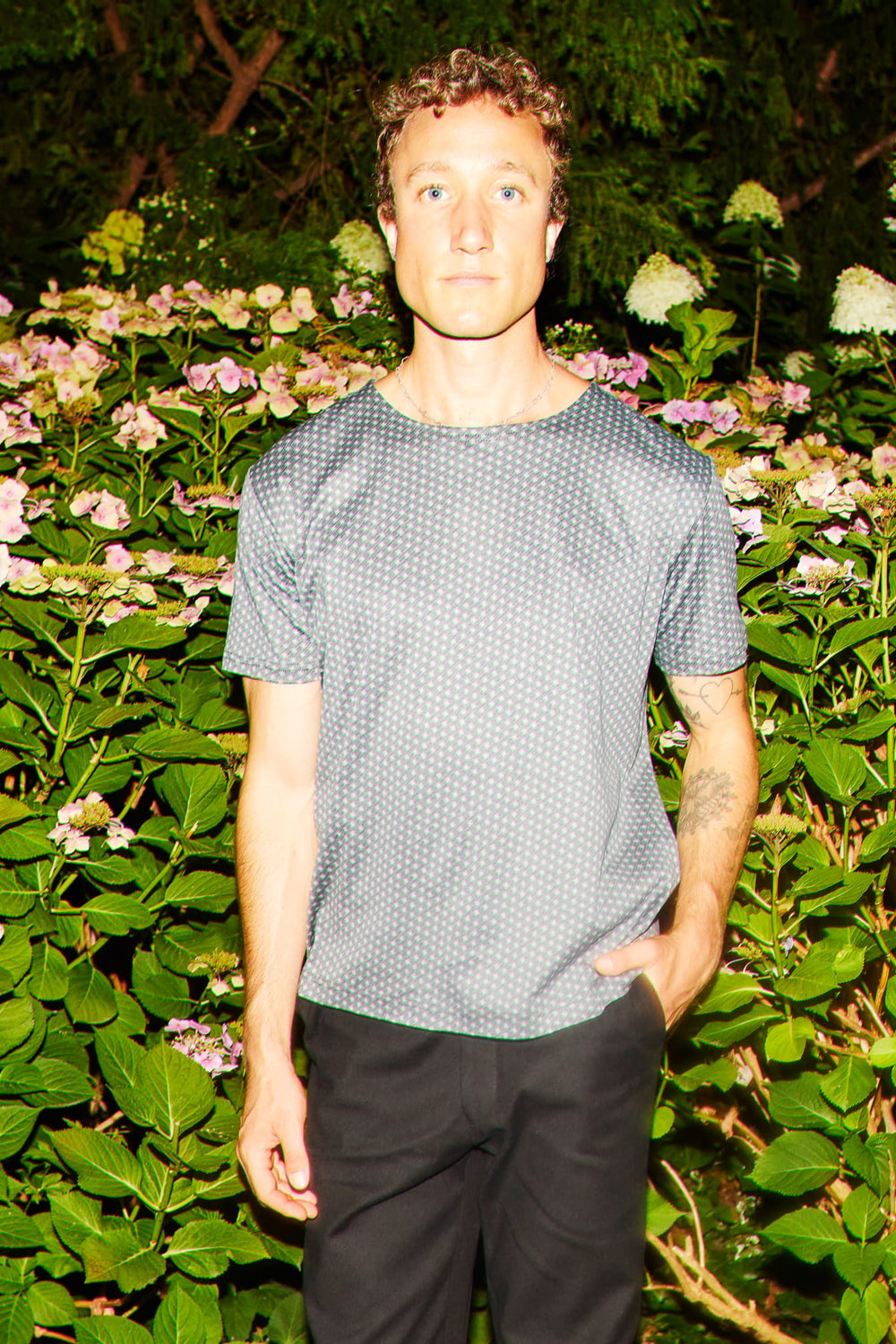 A man wearing a gray short-sleeve t-shirt with a small white geometric pattern and black pants, standing in front of a garden of green foliage and pink flowers.