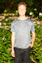 A man wearing a gray short-sleeve t-shirt with a small white geometric pattern and black pants, standing in front of a garden of green foliage and pink flowers.