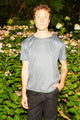 A man wearing a gray short-sleeve t-shirt with a small white geometric pattern and black pants, standing in front of a garden of green foliage and pink flowers.