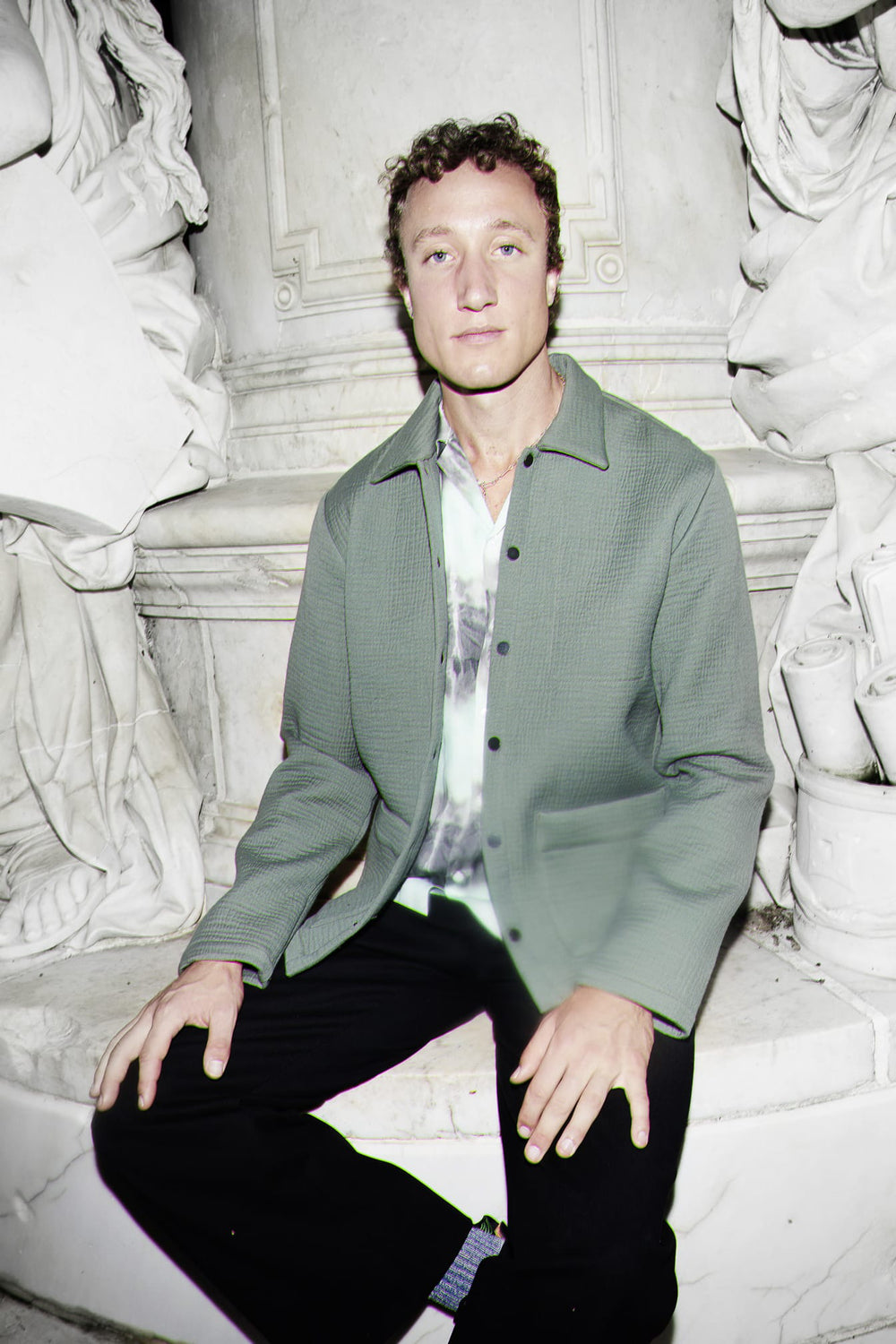 A man wearing a sage green textured workwear overshirt over a white patterned shirt and black trousers, sitting on white stone steps in front of classical statues.