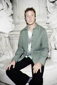 A man wearing a sage green textured workwear overshirt over a white patterned shirt and black trousers, sitting on white stone steps in front of classical statues.